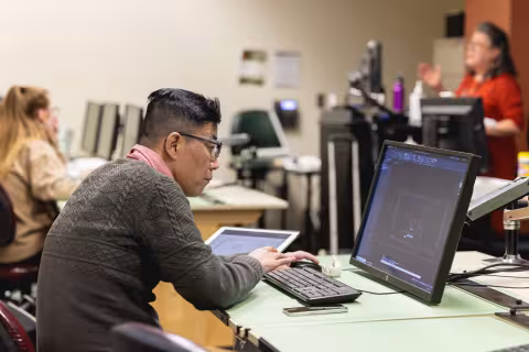 Student looking intently at a computer screen with another student and instructor in the background