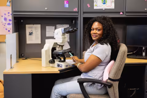 Student at a desk wearing medical scrubs and sitting behind a microscope turned toward the camera and smiling