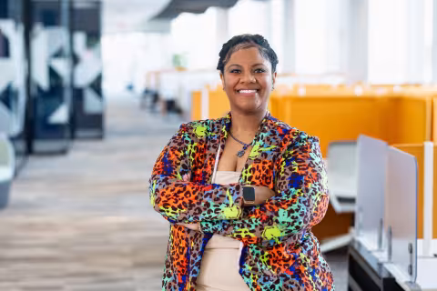 A person stands confidently with arms crossed in a brightly colored leopard-print jacket, smiling warmly in a modern office space.