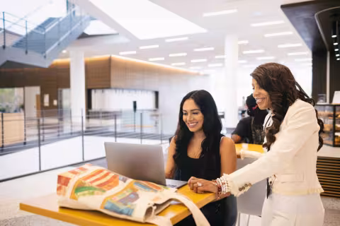 An advisor in a white pant suit standing next to a student who is sitting at a high top table with a laptop in the Parr Center