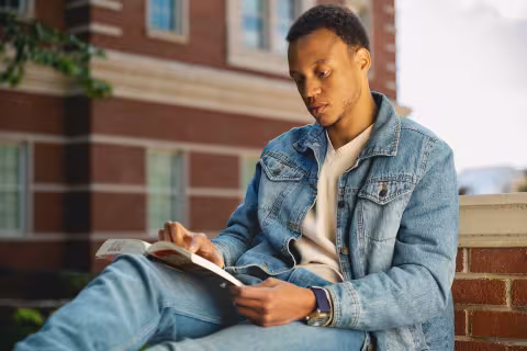 Student reading a book outside, leaning against a brick wall
