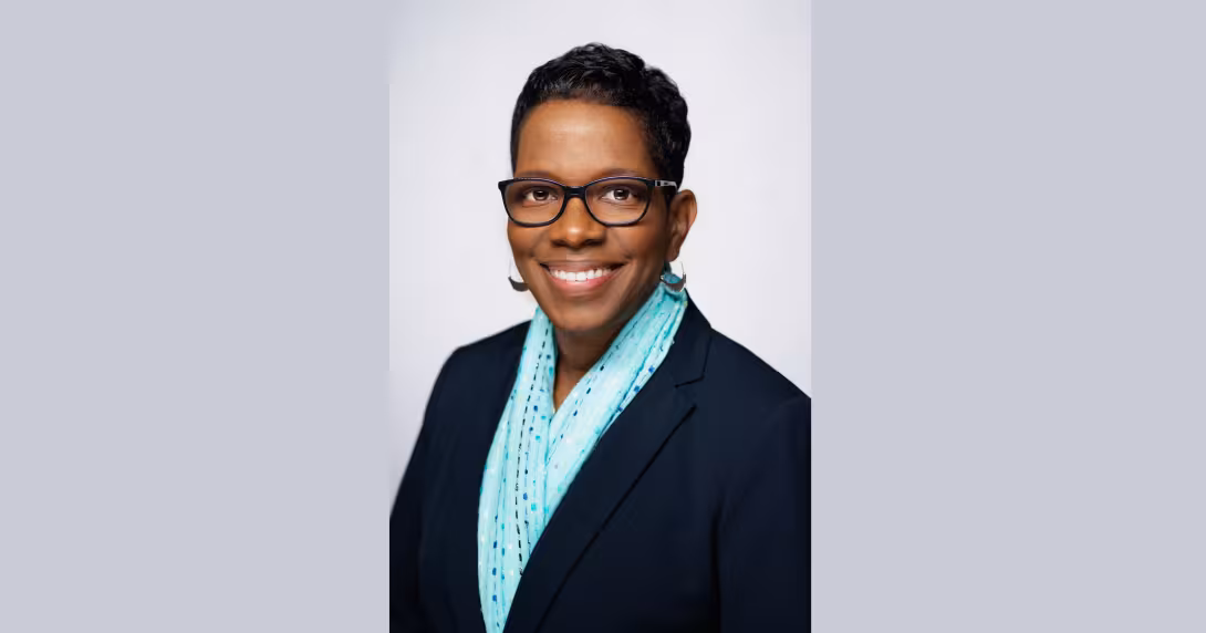 A smiling person with short dark hair, wearing glasses and a blue patterned scarf, dressed in a dark blazer against a plain background.